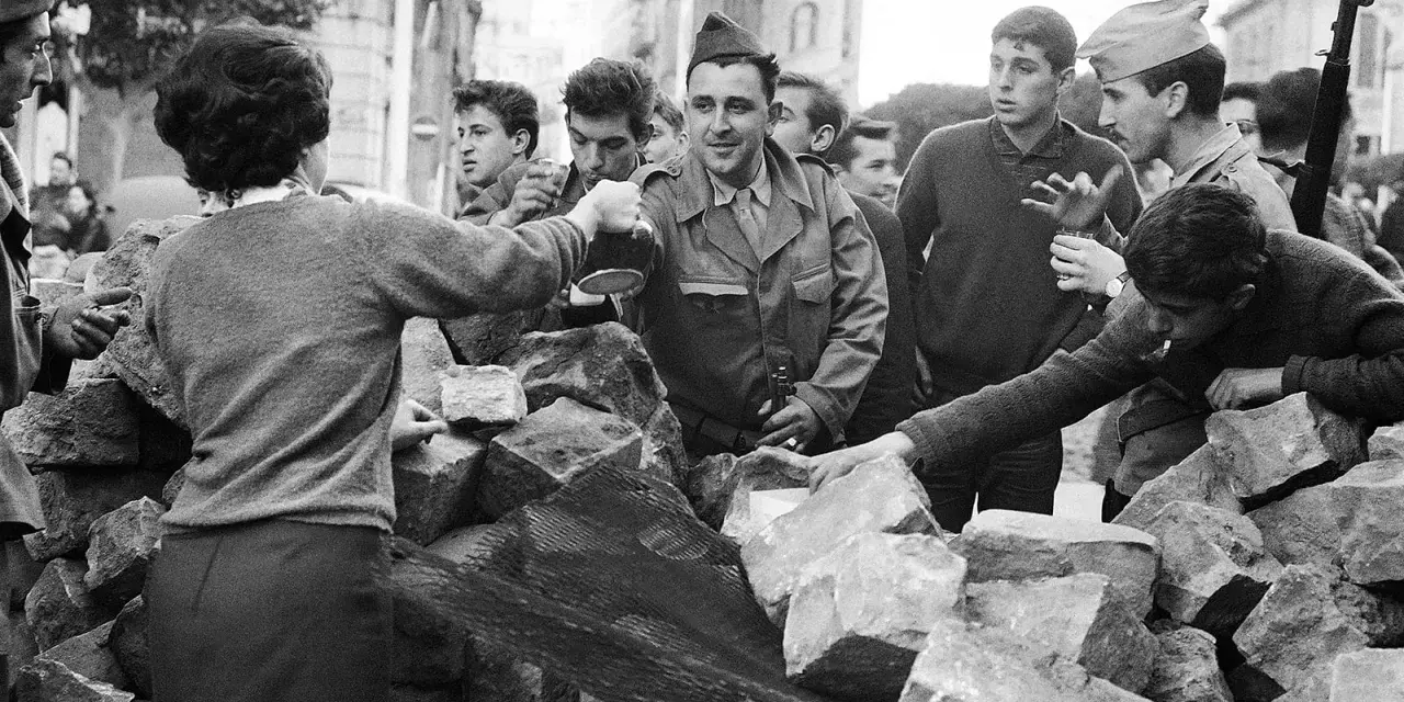 Un grupo de jóvenes y soldados interactúan tras una barricada de piedras en Argelia. Un hombre con uniforme militar sirve bebida.