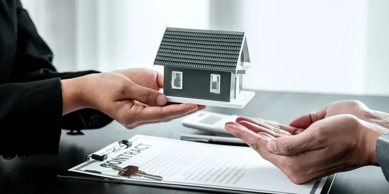 Manos entregan maqueta de casa sobre contrato y llaves. Posible exención IRPF venta vivienda heredada.