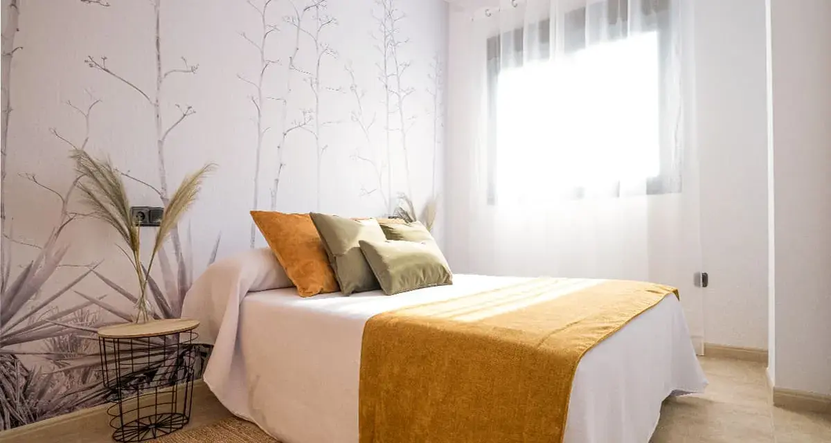 Habitación con papel tapiz de ramas y gotele moderno. Cama con cojines verdes y naranjas, y manta mostaza.