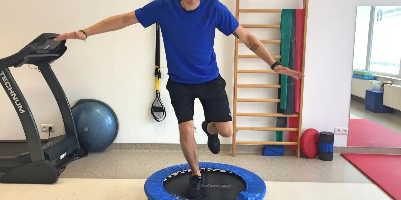 Mężczyzna wykonuje ćwiczenia po rekonstrukcji ACL, balansując na małej trampolinie. W tle bieżnia i drabinka gimnastyczna.