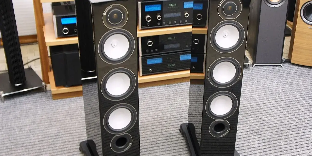 Dwie eleganckie kolumny głośnikowe Monitor Audio RX8 w czarnym połysku, stojące na szarym dywanie, obok sprzętu audio.