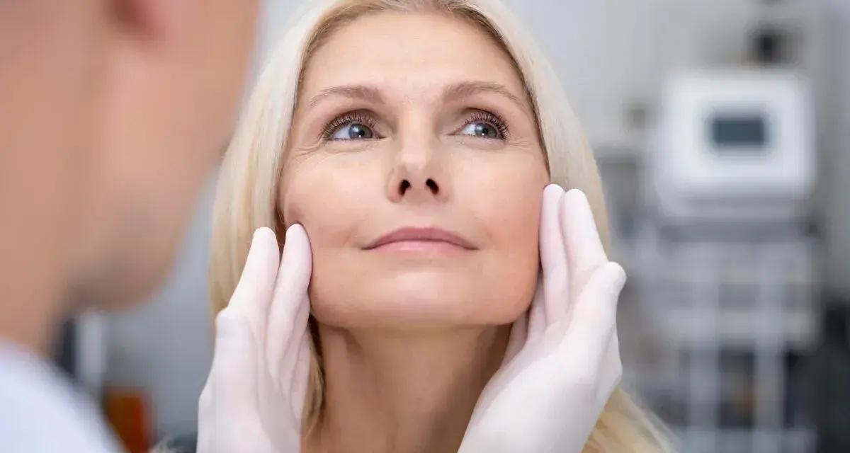 Kobieta z blond włosami poddaje się zabiegowi depilacji laserowej twarzy. Dłonie w rękawiczkach delikatnie dotykają jej policzków.