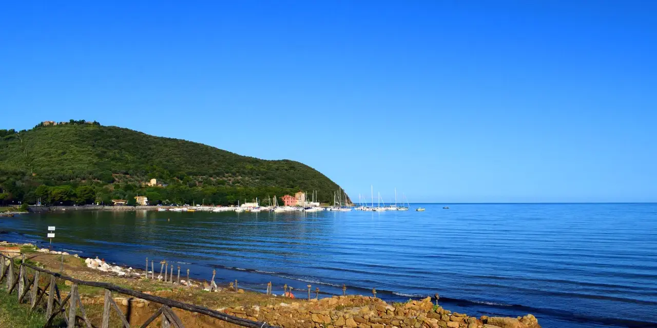 A tranquil bay in Tuscany with sailboats docked, a green hillside, and a rustic wooden fence in the foreground.