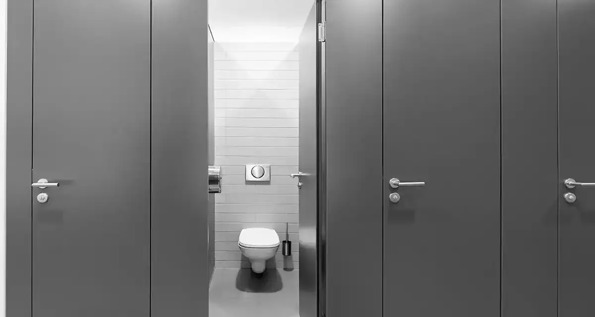 Reihe von grauen Türen zu Toilettenkabinen. Eine Tür ist geöffnet und gibt den Blick auf eine Toilette frei. Gute Frage, wie viele Toiletten pro Mitarbeiter?