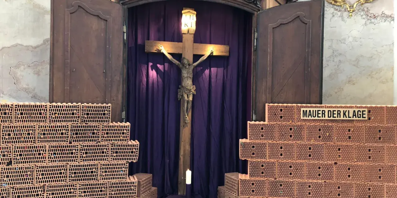 Kreuzweg Stationen: Jesus am Kreuz, umgeben von Ziegelmauern, die die "Mauer der Klage" symbolisieren. Eine Kerze brennt vor dem Kruzifix.