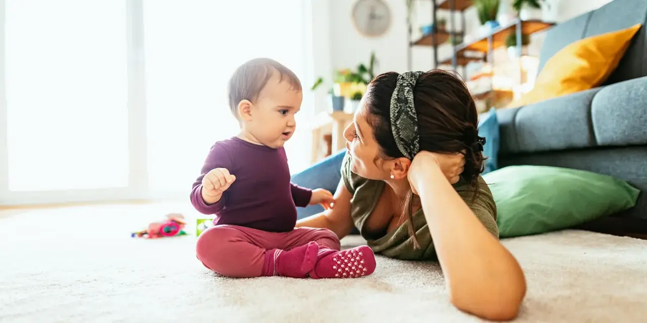 Mama patrzy na dziecko, które siedzi na dywanie. To obrazek pokazujący, co powinien umieć 2 latek: rozwijać się i bawić.