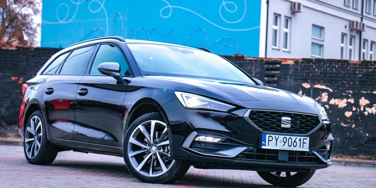 Czarny Seat Leon Sportstourer z polskimi tablicami rejestracyjnymi stoi na brukowanym placu przed budynkiem z niebieską, ozdobną ścianą.