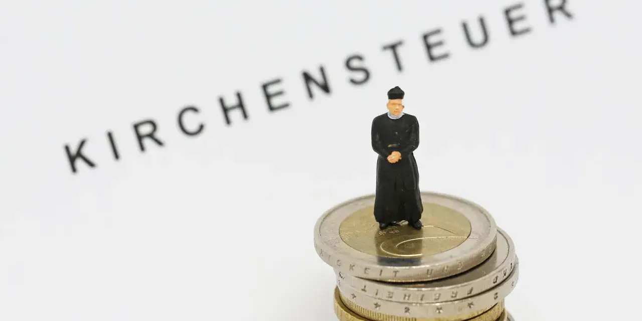 Miniatur-Priester steht auf Münzstapel. Der Schriftzug "Kirchensteuer" im Hintergrund wirft die Frage auf, warum man kirchensteuer zahlt.