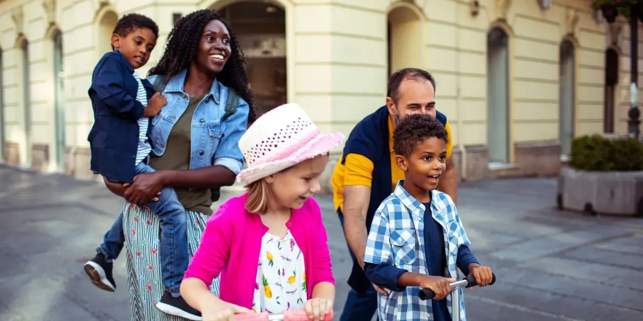 Glückliche Familie unterwegs: Mutter trägt Kind, Vater schiebt Roller. Ein Spagat zwischen Kind und neuem Partner gelingt hier spielerisch.