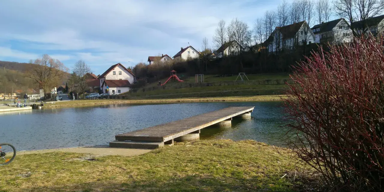 Holzsteg am Badesee Forchheim, mit Häusern im Hintergrund und einem Spielplatz.