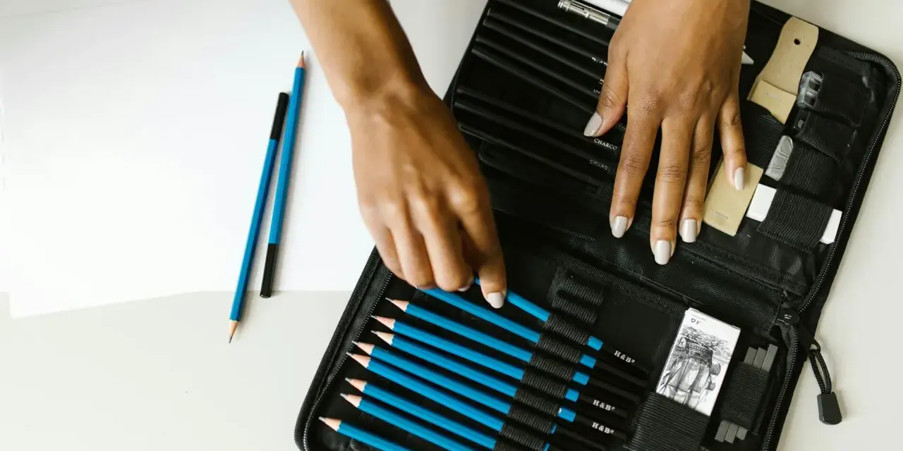 Un étui ouvert révèle une sélection de crayons de différents types pour dessiner, avec des mains choisissant un crayon bleu.