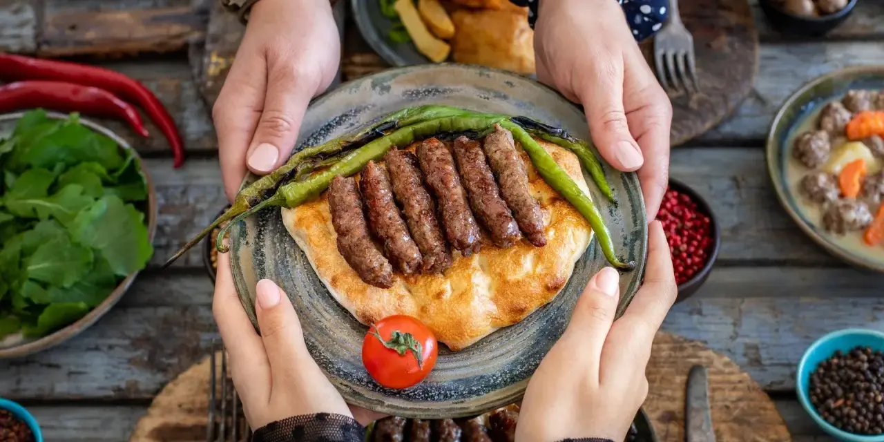 Ręce trzymają talerz z grillowanymi **ćevapi** na płaskim chlebie, ozdobiony papryczkami i pomidorem. Obok sałatka i inne dania.