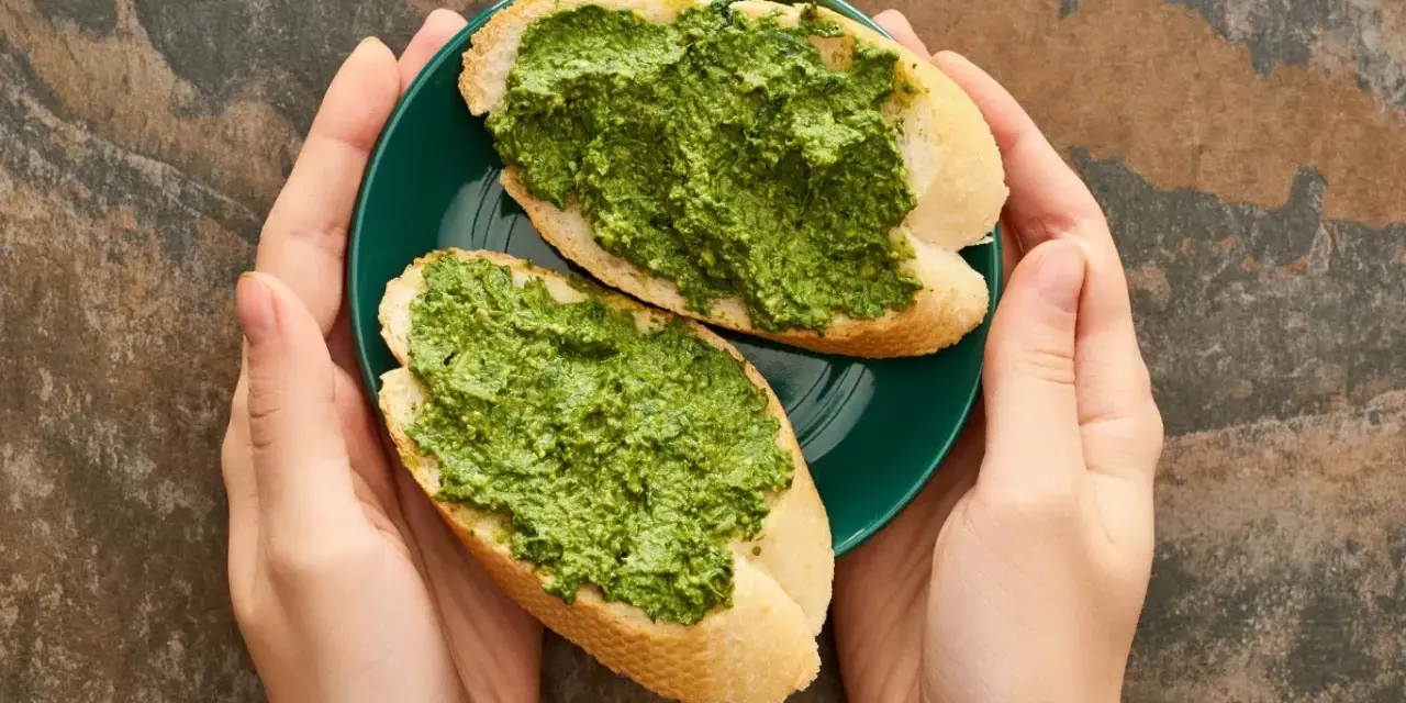 Dwie kromki bagietki z aromatycznym pesto z pietruszki na zielonym talerzyku, trzymanym w dłoniach.