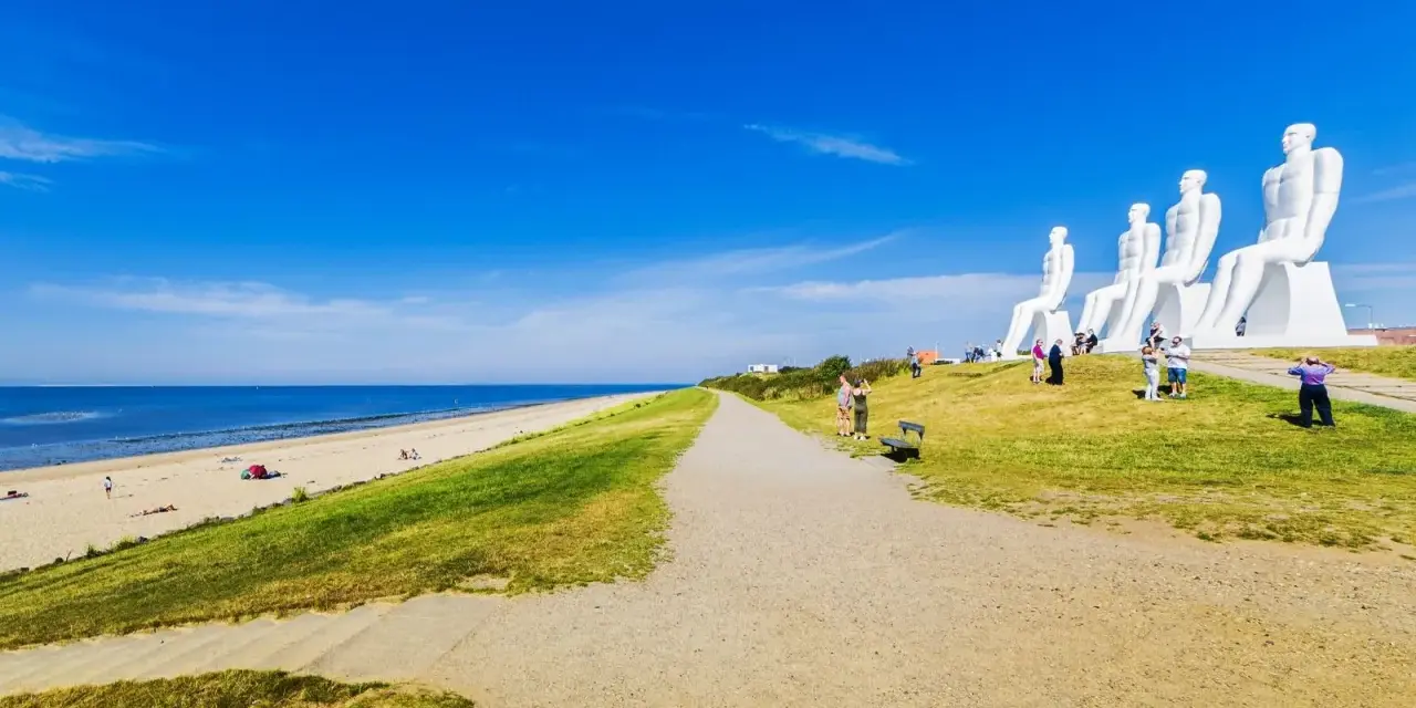 De vita sandstränderna i Danmark möter en grönskande sluttning med gigantiska sittande figurer. En grusväg leder mot havet under en klarblå himmel.