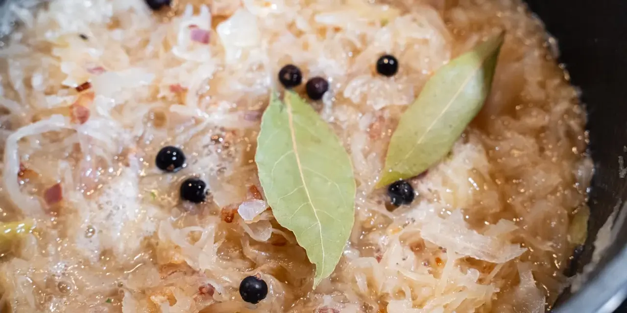 Sauerkraut köchelt mit Lorbeerblättern und Wacholderbeeren, um den Geschmack zu verfeinern. Kleine rote Zwiebelstücke sind ebenfalls sichtbar.