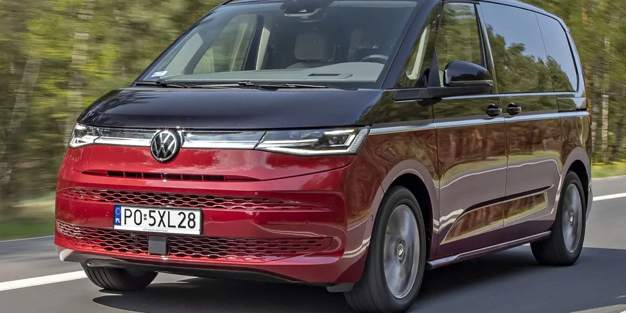 Nowy Volkswagen van w kolorze czerwono-czarnym pędzi po drodze, otoczony zielenią drzew.