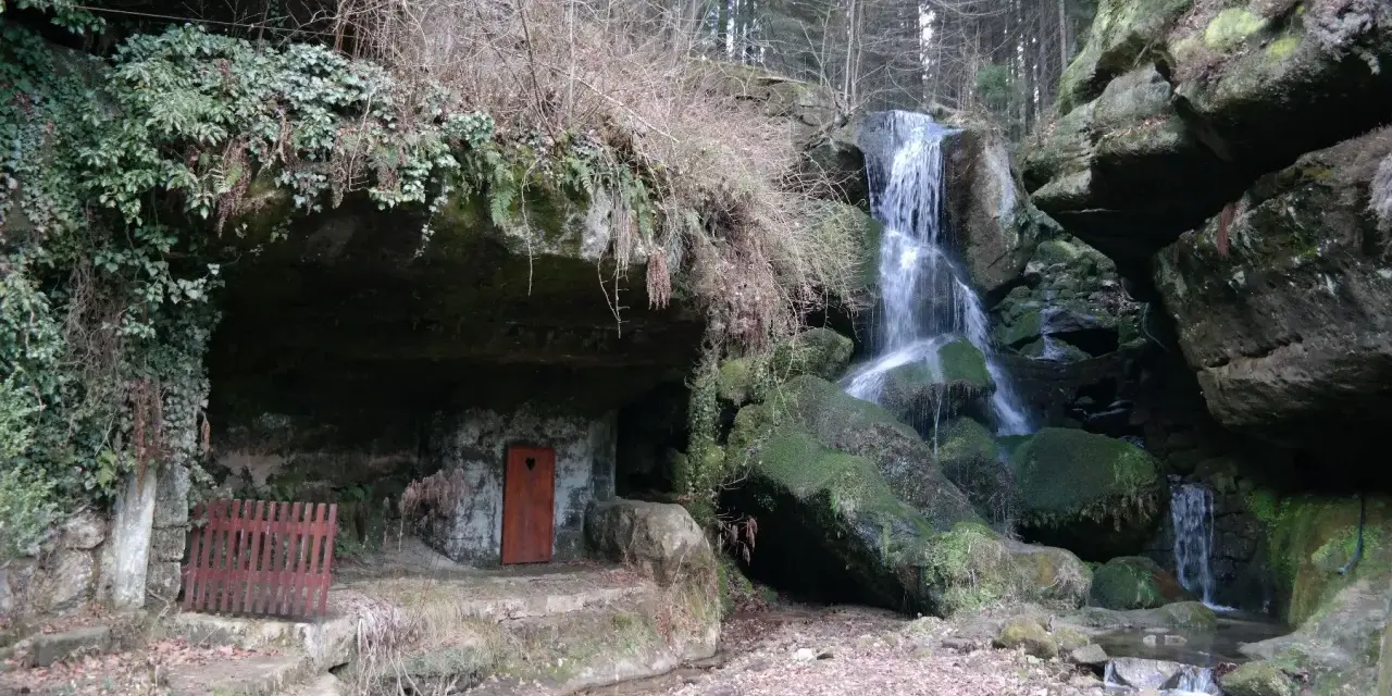 Ein versteckter Wasserfall bei Bad Schandau, mit einer kleinen Höhle und einer roten Tür.