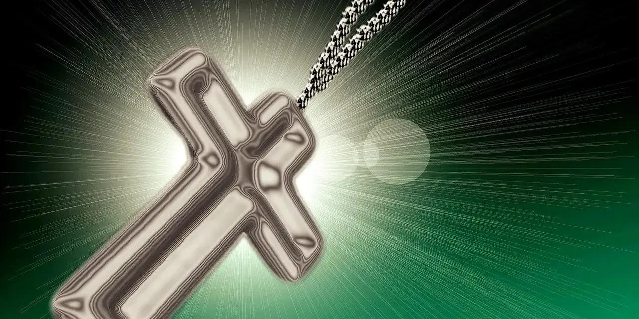 Metalowy krzyż na sznurku, otoczony blaskiem. Symbolizuje nadzieję i nawrócenie, światło rozchodzące się na zielonym tle.