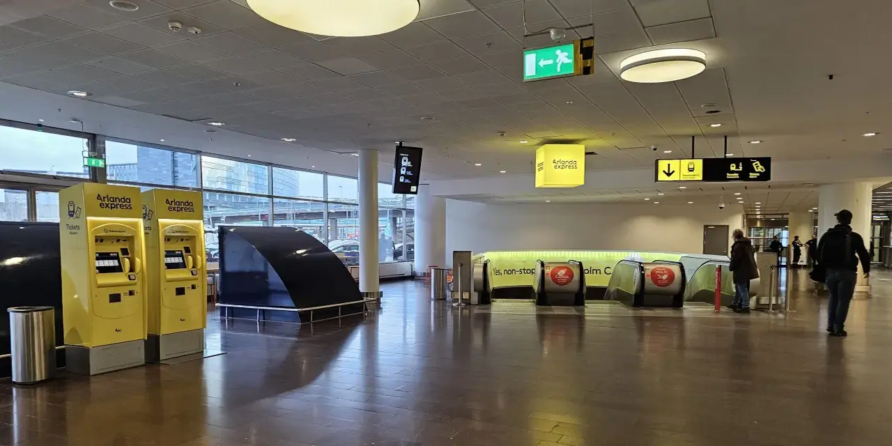 Żółte automaty biletowe Arlanda Express na lotnisku w Sztokholmie. W tle ruchome schody i znaki kierujące.
