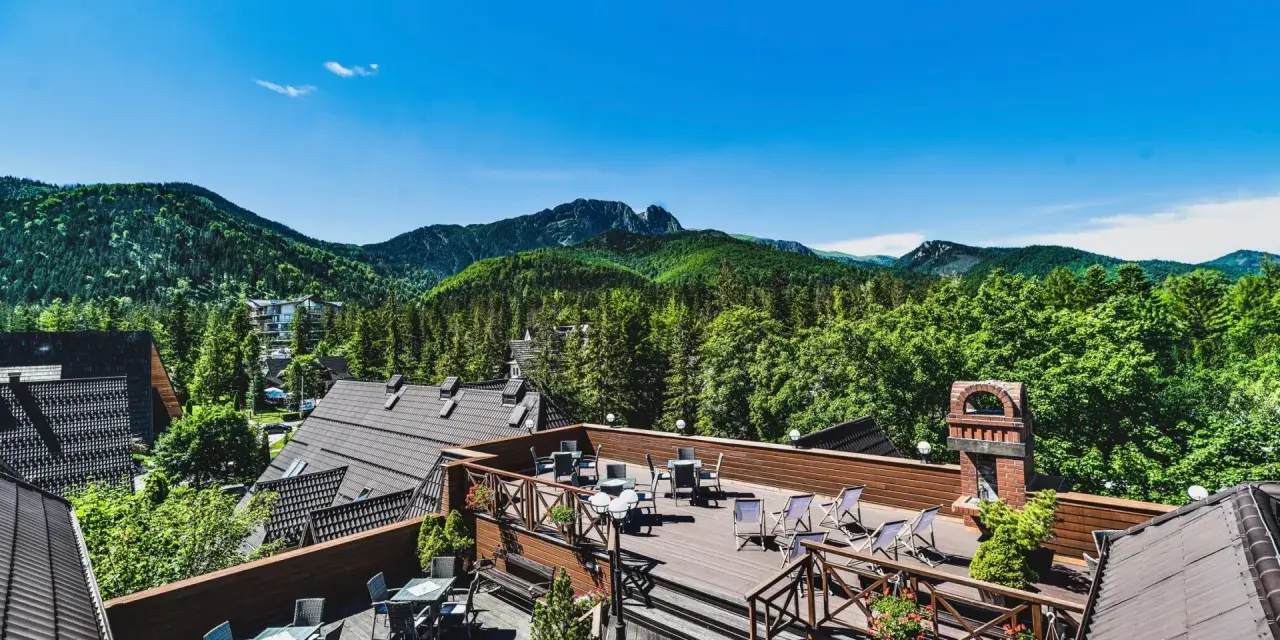 Hotel z widokiem na góry. Taras z meblami i leżakami, otoczony zielenią i górami.