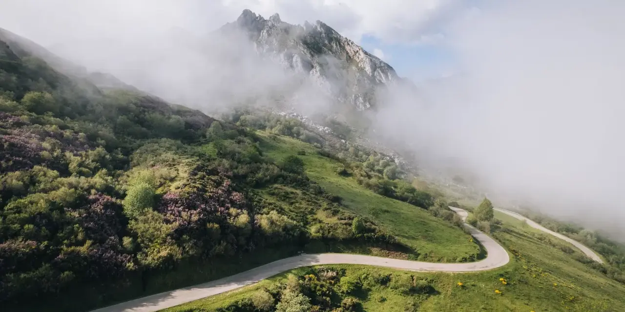 Strada tortuosa che sale verso le vette nebbiose, evocando l'ascesa al **Col du Tourmalet**.