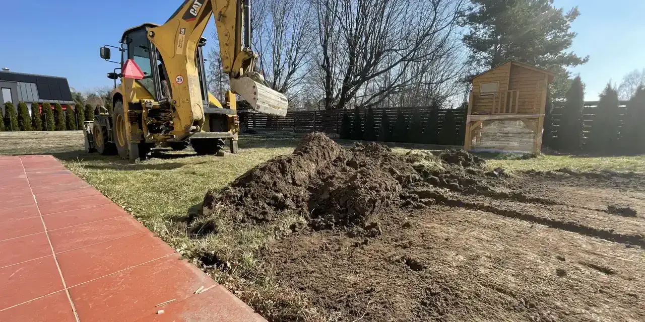 Koparka wykonuje niwelację terenu, przesuwając ziemię. W tle widać domek na drzewie i ogrodzenie.