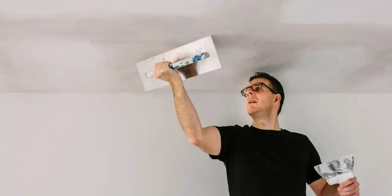 Mężczyzna w okularach wykonuje tynkowanie sufitu, używając kielni i packi.