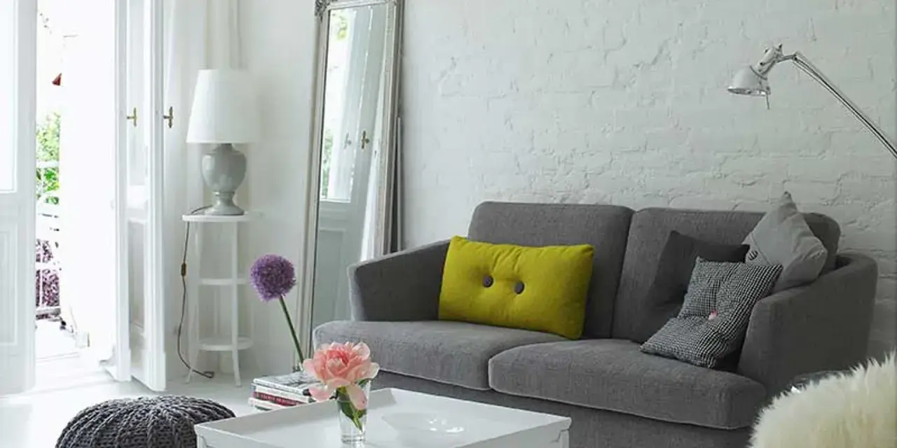 Sofá gris con cojines, espejo alto y lámpara. Trucos para decorar espacios pequeños con estilo y amplitud.