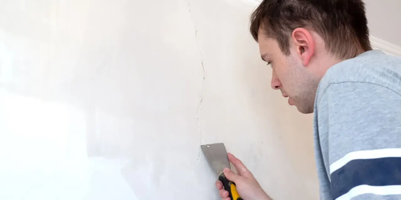 Uomo con spatola ripara una crepa su un muro bianco.