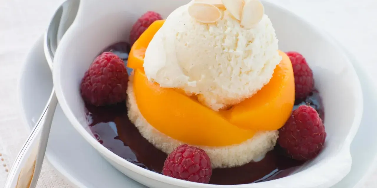 Ein köstlicher Pfirsich Melba: Vanilleeis auf Pfirsichspalten, umgeben von Himbeeren und dunkler Sauce, garniert mit Mandelblättchen.