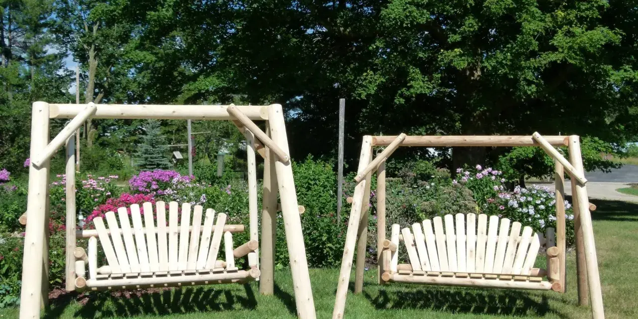 Two wooden porch swings stand on a grassy lawn, surrounded by colorful flowers and lush trees. This image offers inspiration for how to hang a porch swing.
