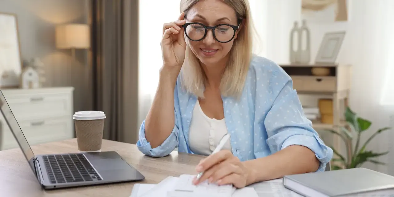 Femme aux lunettes calculant, laptop et café à proximité. Elle semble gérer des finances, peut-être une réduction de capital non motivée par des pertes.