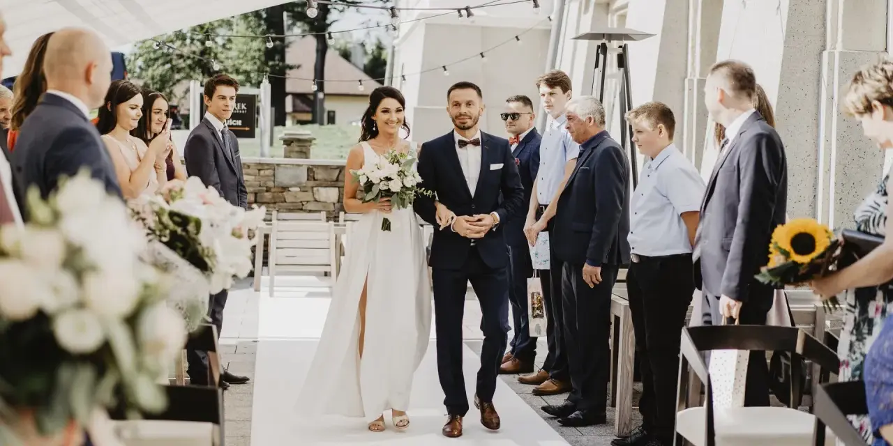 Para idzie do ołtarza, składając przysięgę małżeńską cywilną. Panna młoda w białej sukni, pan młody w granatowym garniturze.