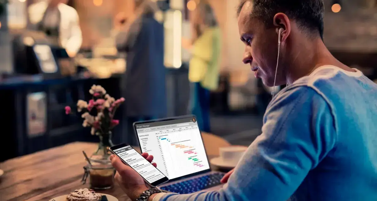 Hombre revisa datos de business intelligence hostelería en su laptop y móvil, sentado en una cafetería.