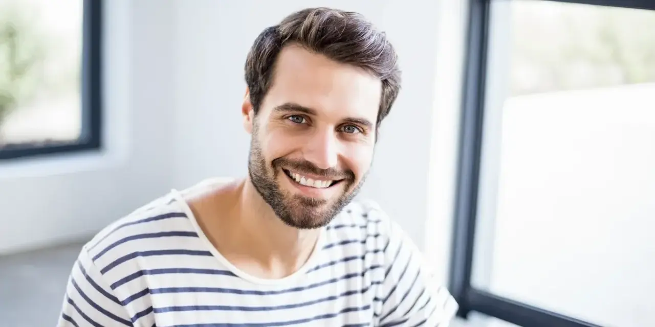 Uśmiechnięty mężczyzna o imieniu Marcin, z brodą i w pasiastym t-shircie, emanuje pozytywną energią. Znaczenie imienia Marcin to wojownik.