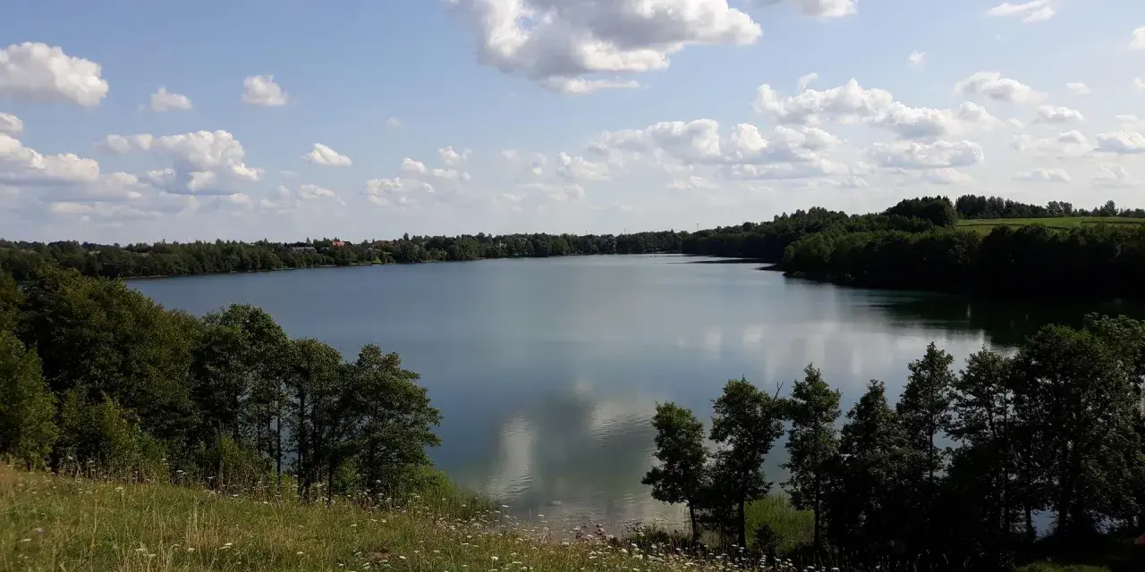 Spokojne jezioro Hańcza otoczone zielonymi lasami, z białymi chmurami na błękitnym niebie.