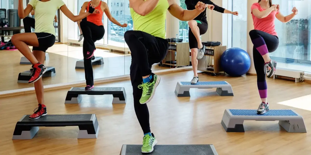 En grupp människor utför step ups i ett gym. De har roligt medan de tränar.