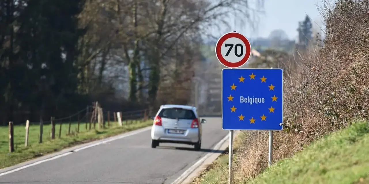 Znak graniczny Belgii, gdzie potrzebny paszport, na tle drogi i jadącego samochodu.