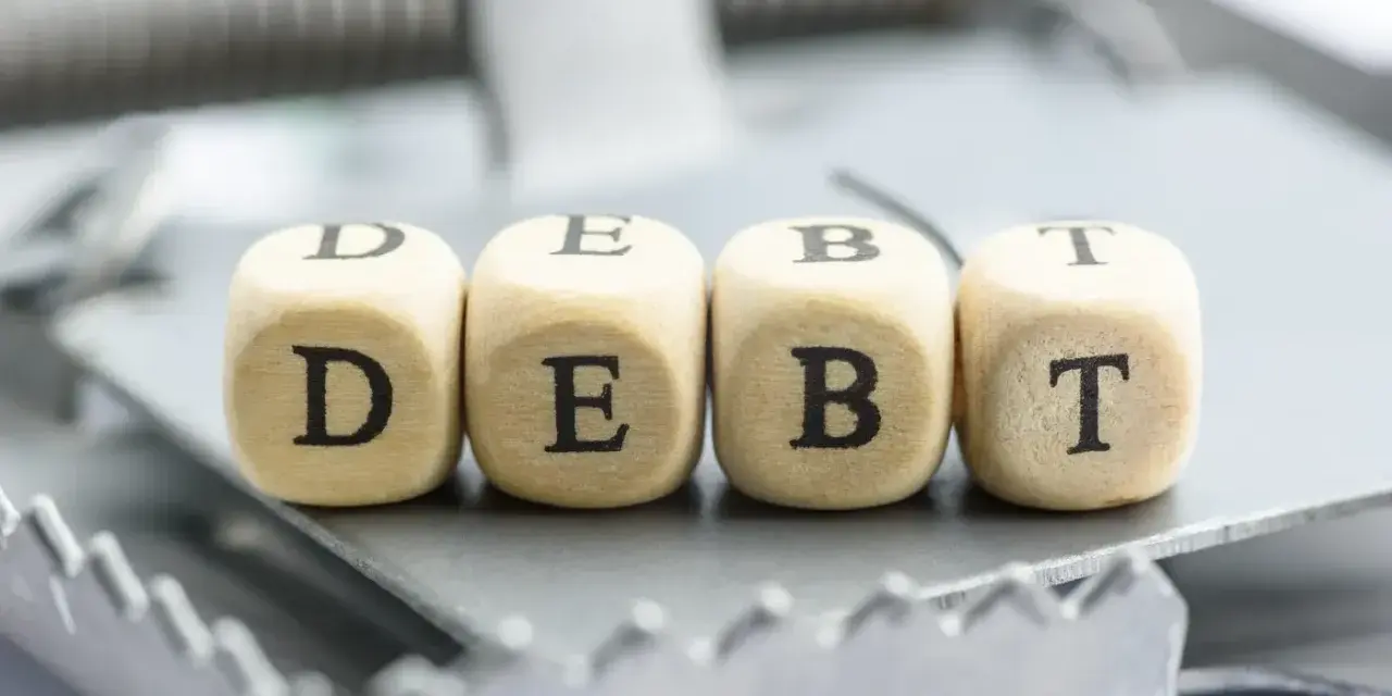 Cztery drewniane kostki z literami D, E, B, T tworzą słowo "DEBT" na pułapce. To symbol finansowych zobowiązań, które mogą prowadzić do trudnej sytuacji, jak datio in solutum.