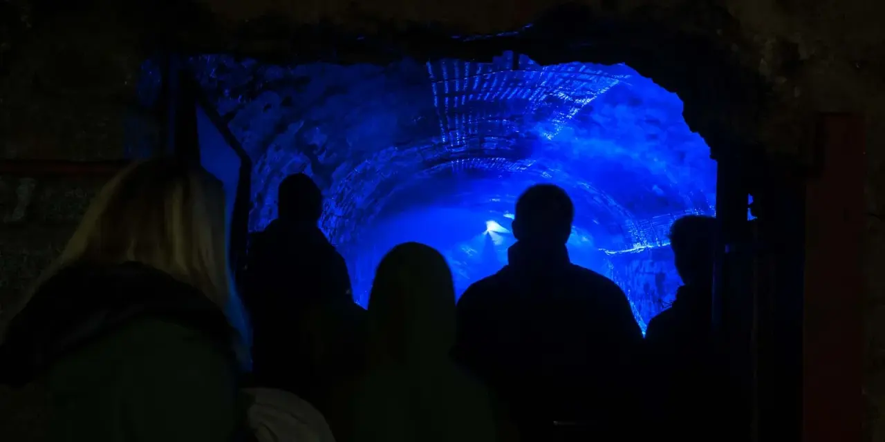 Grupa turystów wchodzi do tunelu oświetlonego niebieskim światłem. To część podziemnej trasy turystycznej w dawnej kopalni uranu Kowary.