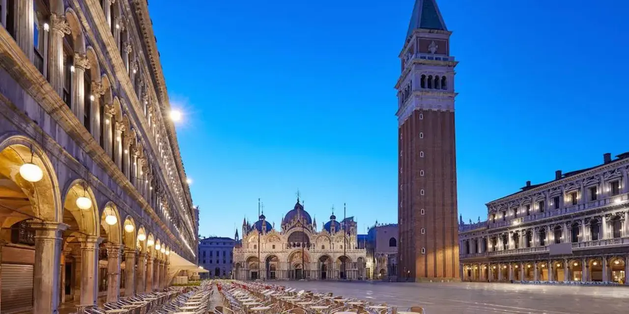 Piazza San Marco a Venezia, con la Basilica e il Campanile, testimoni della ricca storia della città. Tavolini all'aperto creano un'atmosfera serale.