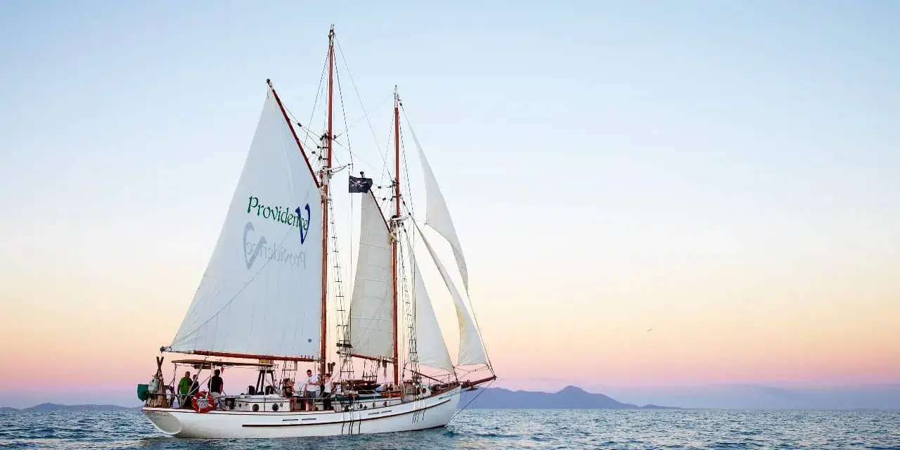 Żaglowiec "Providence" płynie po spokojnych wodach, zbliżając się do Whitehaven Beach. Zachód słońca maluje niebo pastelowymi barwami.