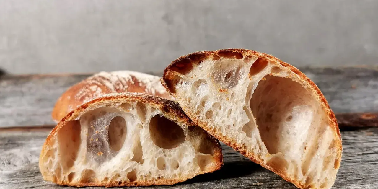 Zwei Hälften eines rustikalen ciabatta mit sauerteig, mit großer, luftiger Krume und knuspriger Kruste, liegen auf einem Holzbrett.