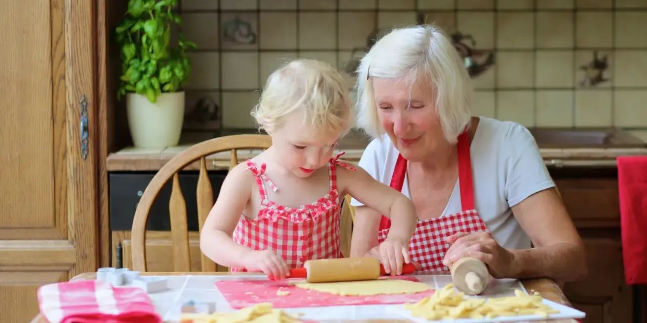 Babcia z wnuczką pieką ciasteczka. Dziewczynka wałkuje ciasto, a babcia z uśmiechem patrzy na jej pracę.