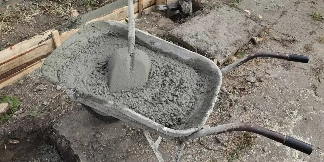 Taczka z betonem i łopatą czeka na użycie przy budowie fundamentów. W tle widać szalunki i przygotowane zbrojenie.