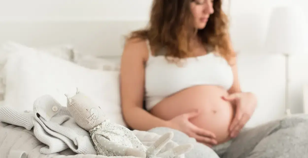 Czekając na pierwsze kopnięcia, przyszła mama głaszcze brzuszek. Obok leżą ubranka i przytulanka dla maluszka.