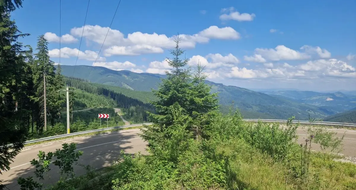 Górzysty krajobraz Rumunii z krętą drogą i lasami. W oddali widać pasma górskie, a na niebie chmury.