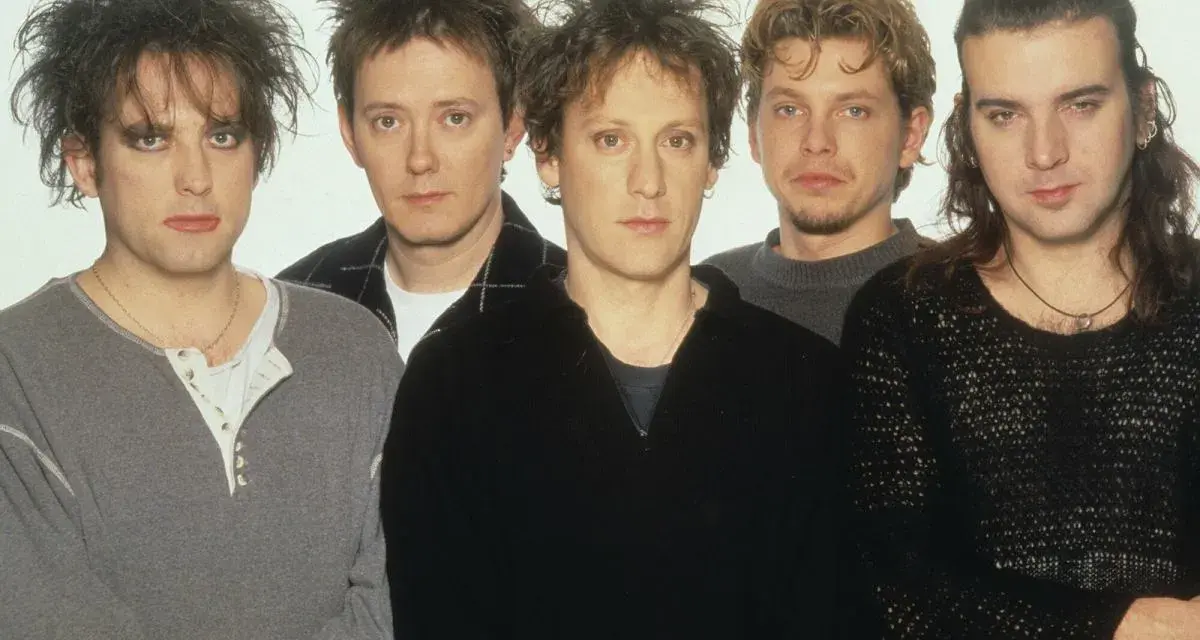 Pięciu członków zespołu The Cure pozuje do zdjęcia. Robert Smith z charakterystyczną fryzurą i makijażem na pierwszym planie.