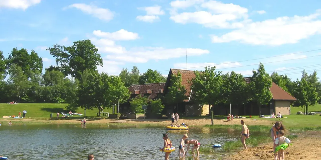 Sonniger Tag am badesee fredenbeck. Kinder planschen im Wasser und am Sandstrand, während im Hintergrund Gebäude und Bäume zu sehen sind.