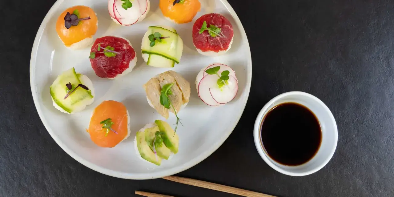 Na talerzu ułożone są kolorowe kulki sushi z łososiem, wołowiną, awokado i rzodkiewką. Obok miseczka sosu sojowego i pałeczki.